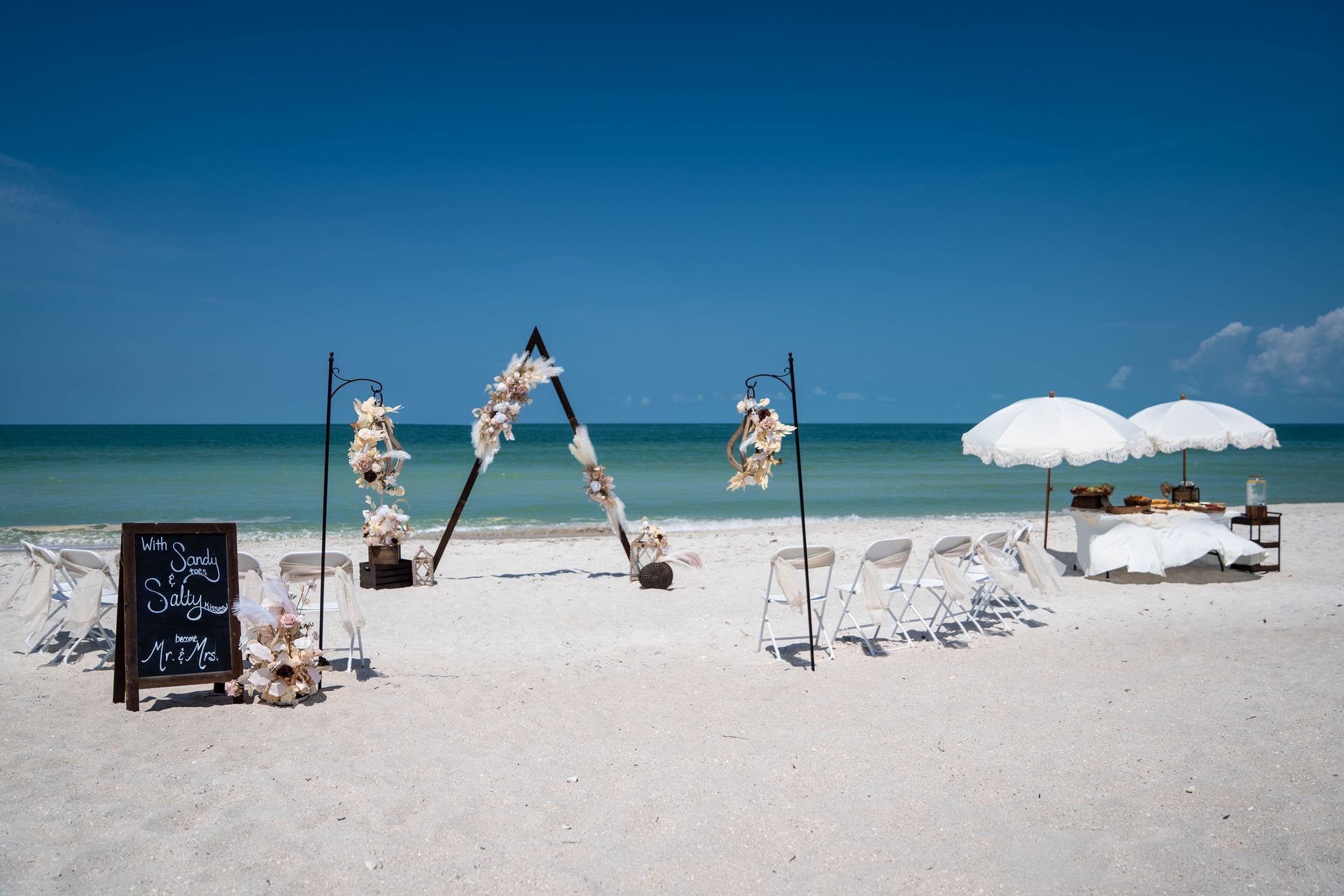 Beach wedding setup with arch, chairs, sign, and umbrellas on a sunny day.