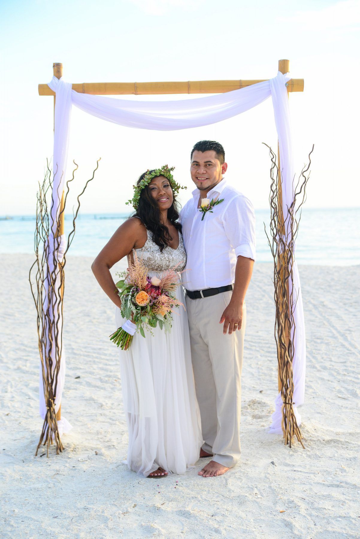 Bride and groom posing on beach under a decorated archway. Bride holds flowers, groom wears button-down shirt.