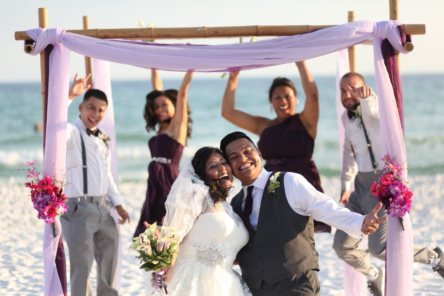 Newlyweds and wedding party celebrating on a beach under a draped bamboo arch.