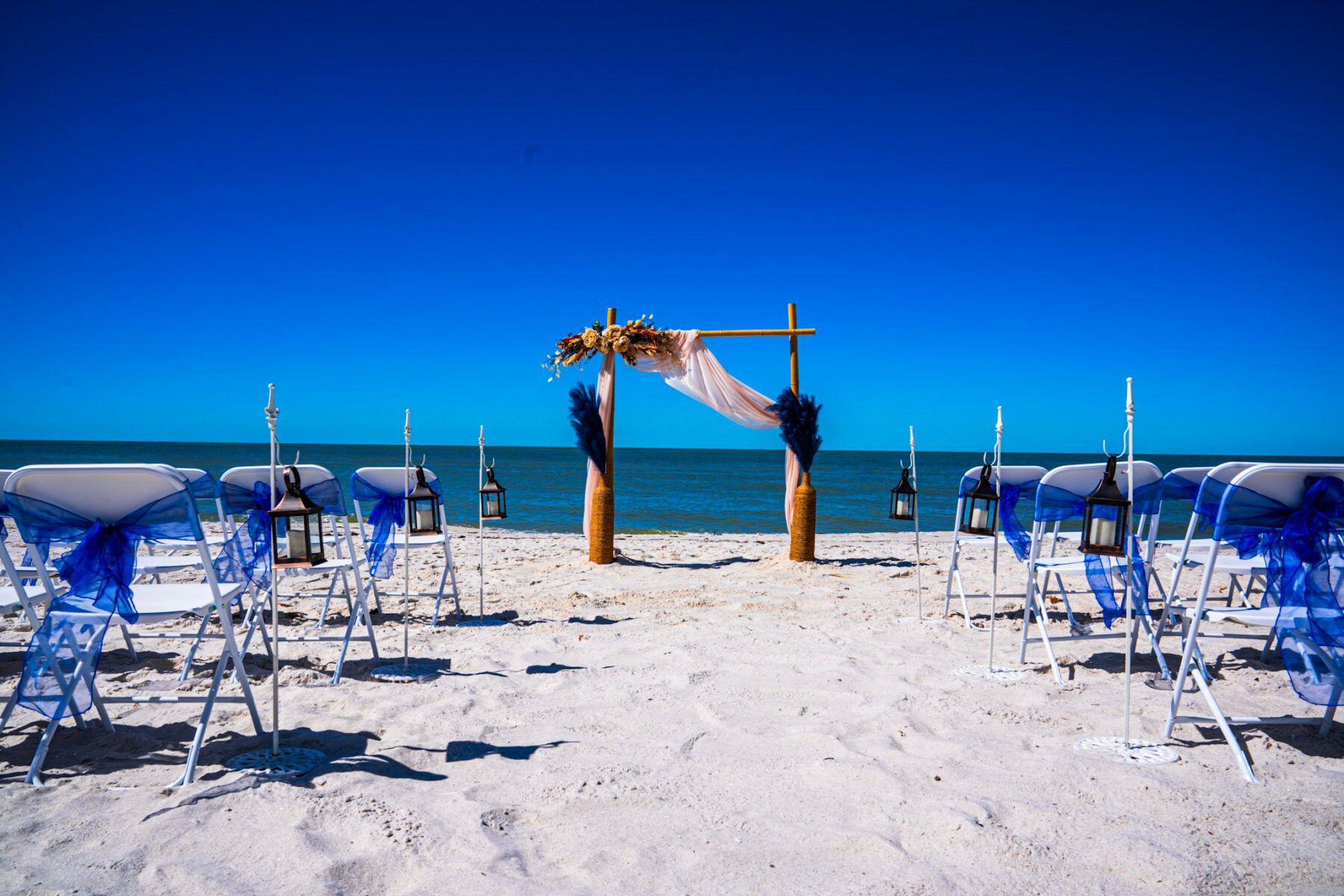 Beach wedding ceremony setup with white chairs, arch, and blue accents.