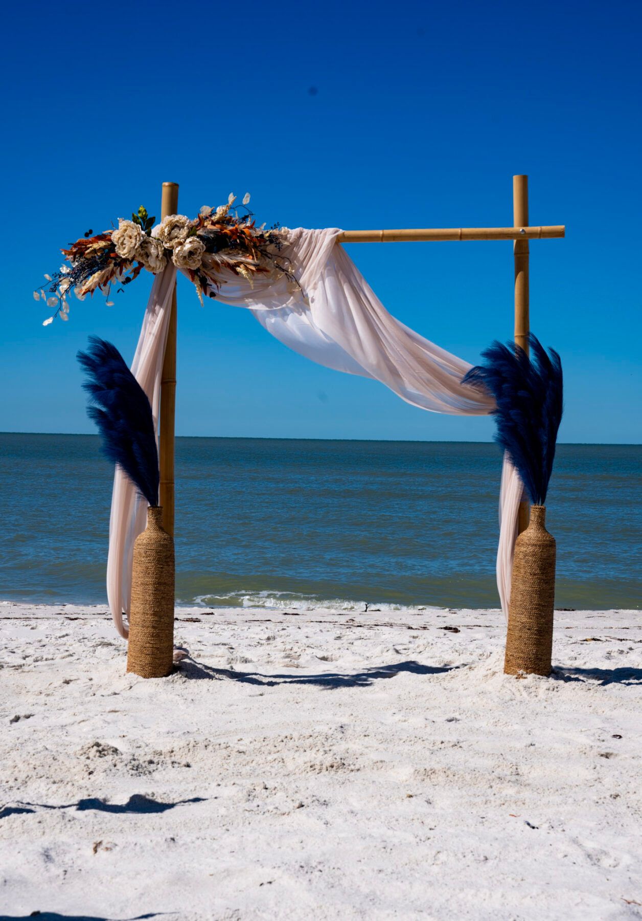 Wedding arch on beach, decorated with flowers, blue plumes, and white fabric, against a blue sky.