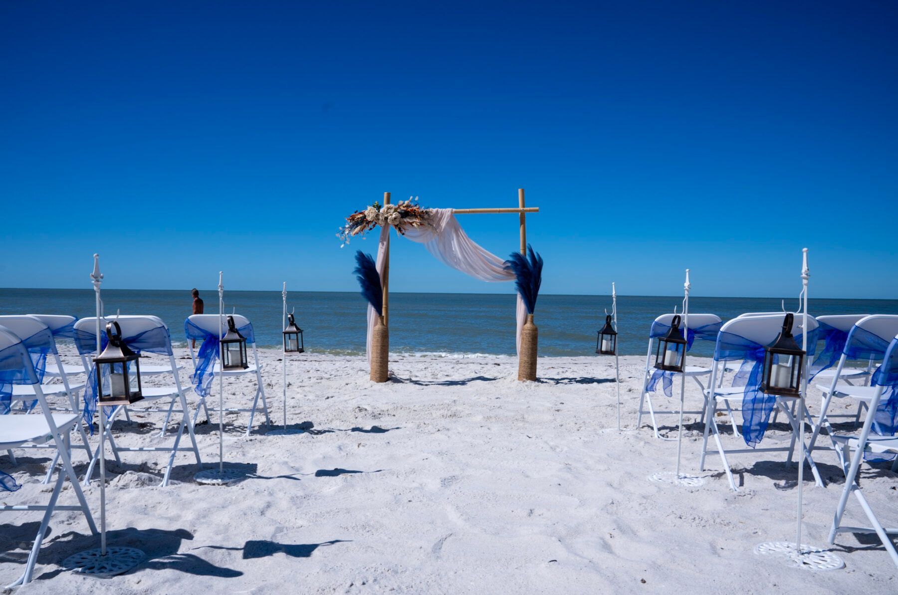 Beach wedding setup with chairs, arch, and lanterns against a blue sky and ocean.