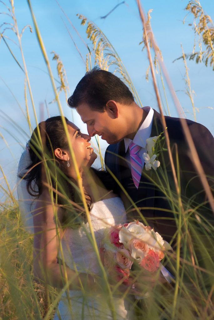 Bride and groom embrace, touching noses in field of tall grass with blue sky backdrop.
