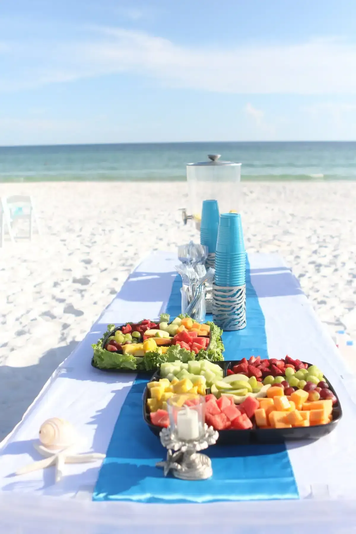 Beachside table with fruit platters, drinks, and blue decor.