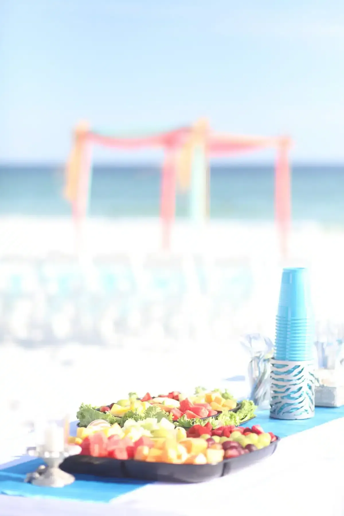 Beachside buffet with fruit salad, blue cups, and an arch, with the ocean in the background.