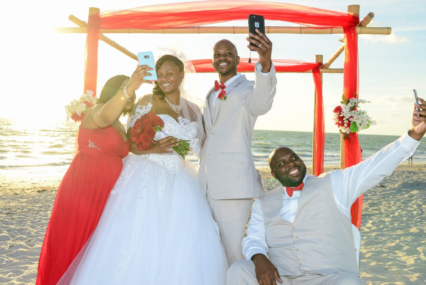 Wedding group taking selfies on a beach. Bride and groom with two others under a red canopy.