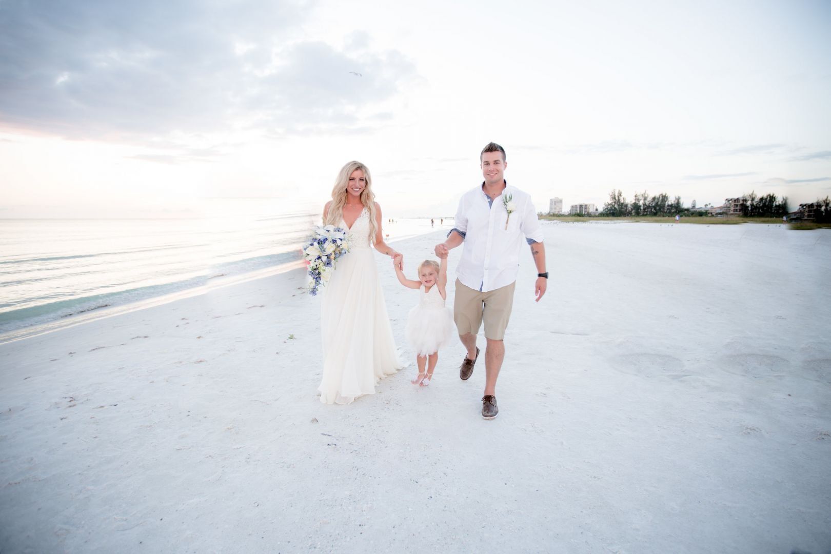 Family walks on a beach after a wedding: woman in a gown, man in shorts, child in white dress.