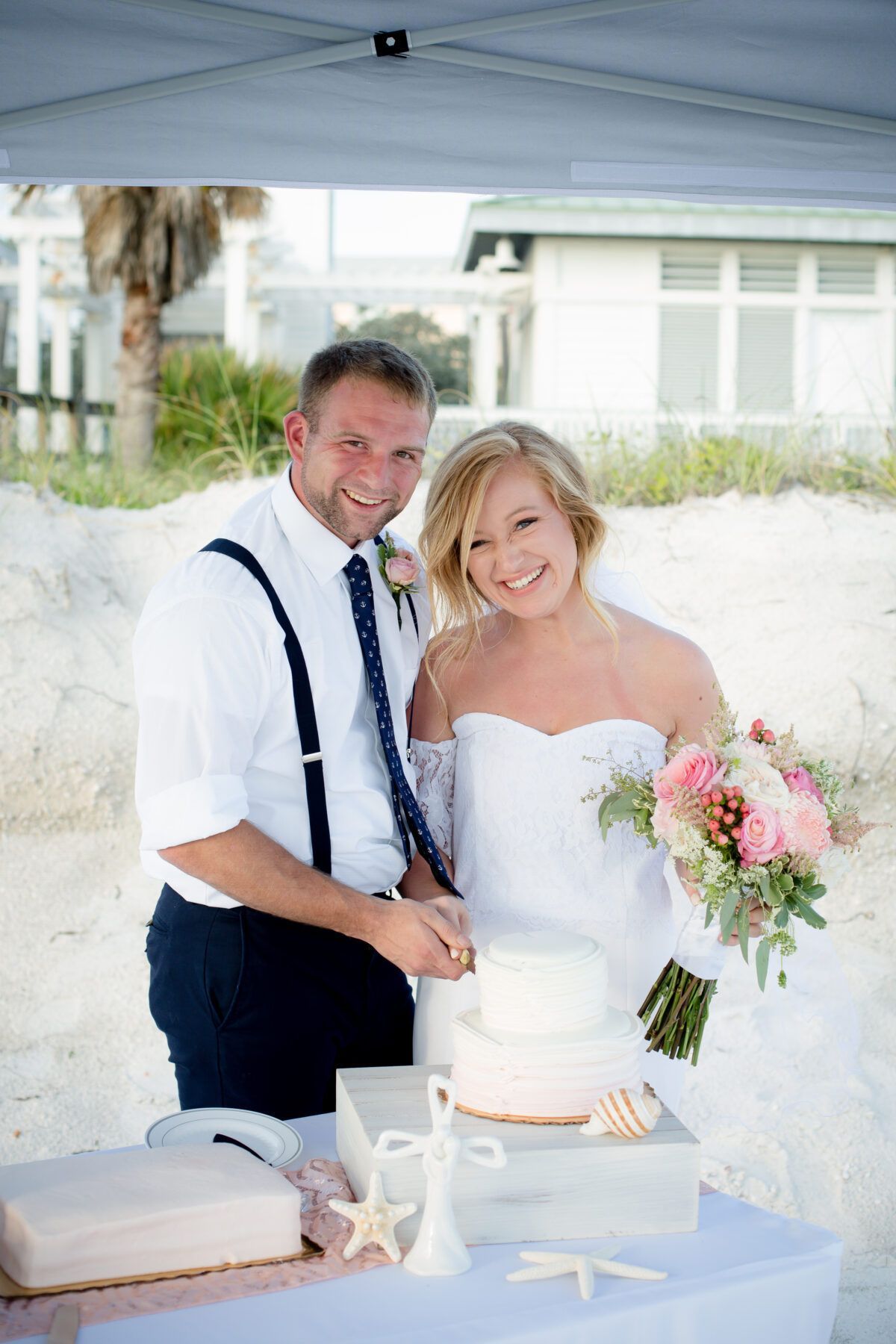 Bride and groom cut wedding cake on the beach under a canopy; she holds flowers, he smiles, they wear formal attire.
