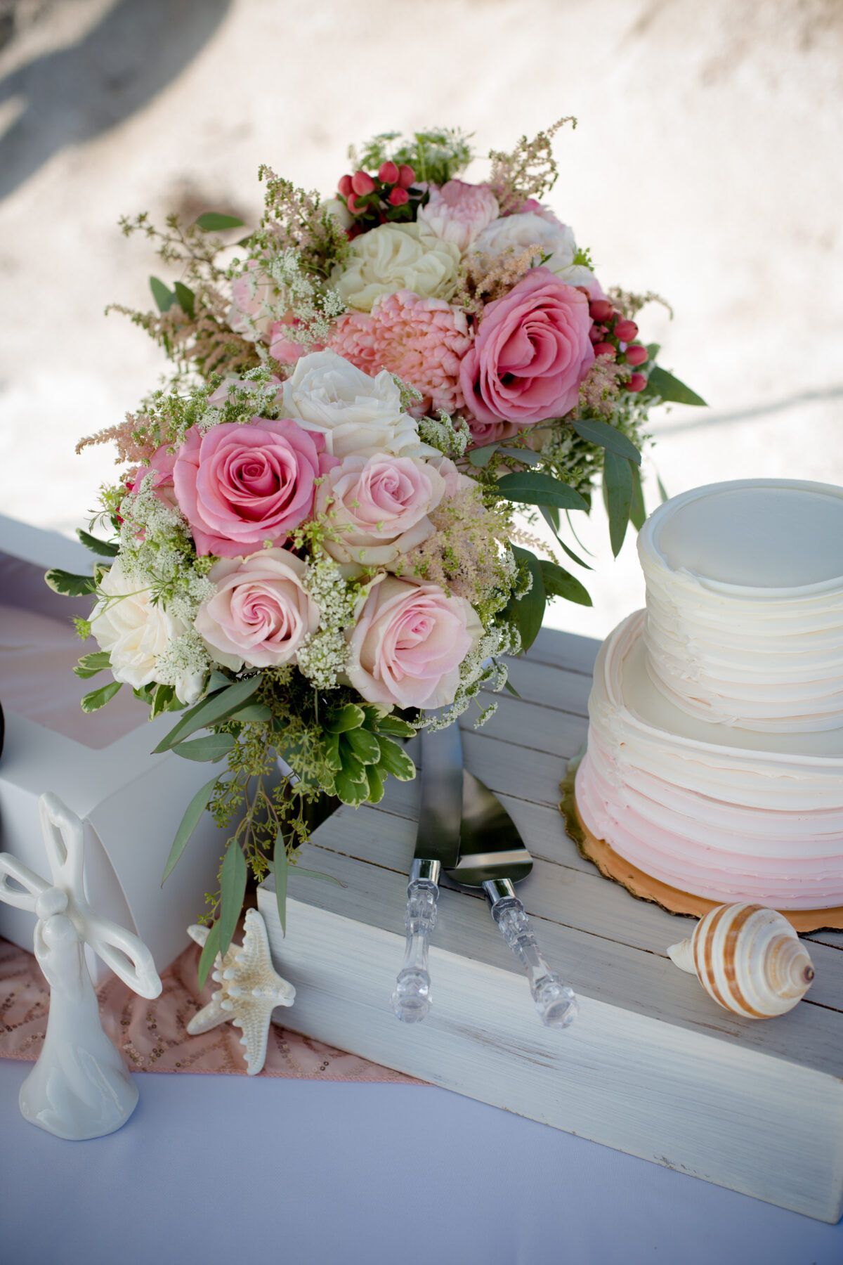 Wedding bouquet with pink roses, cake, and seashell on a beach setting.