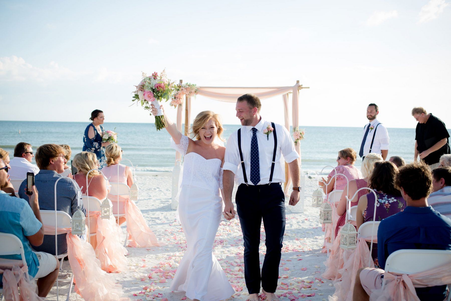 Bride and groom exiting beach wedding ceremony; bride raises arm, holding bouquet; ocean in background.