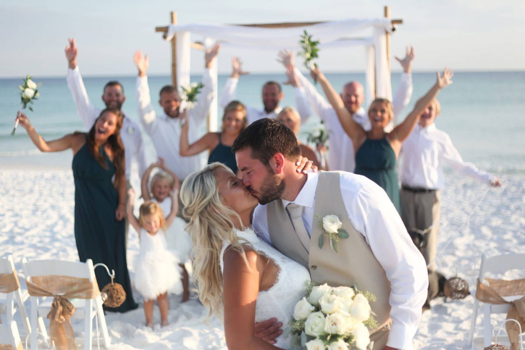 Bride and groom kissing on a beach; wedding party cheering in the background.