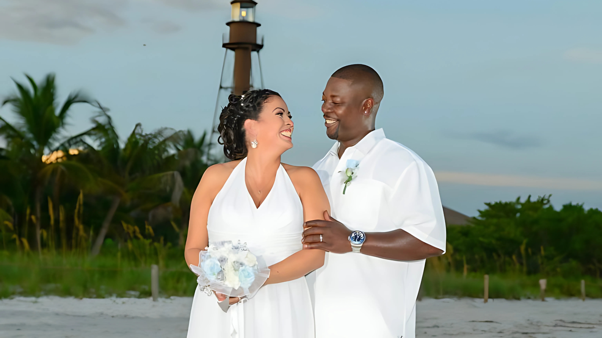 Couple smiles at each other on a beach, holding a bouquet, with a lighthouse in the background.