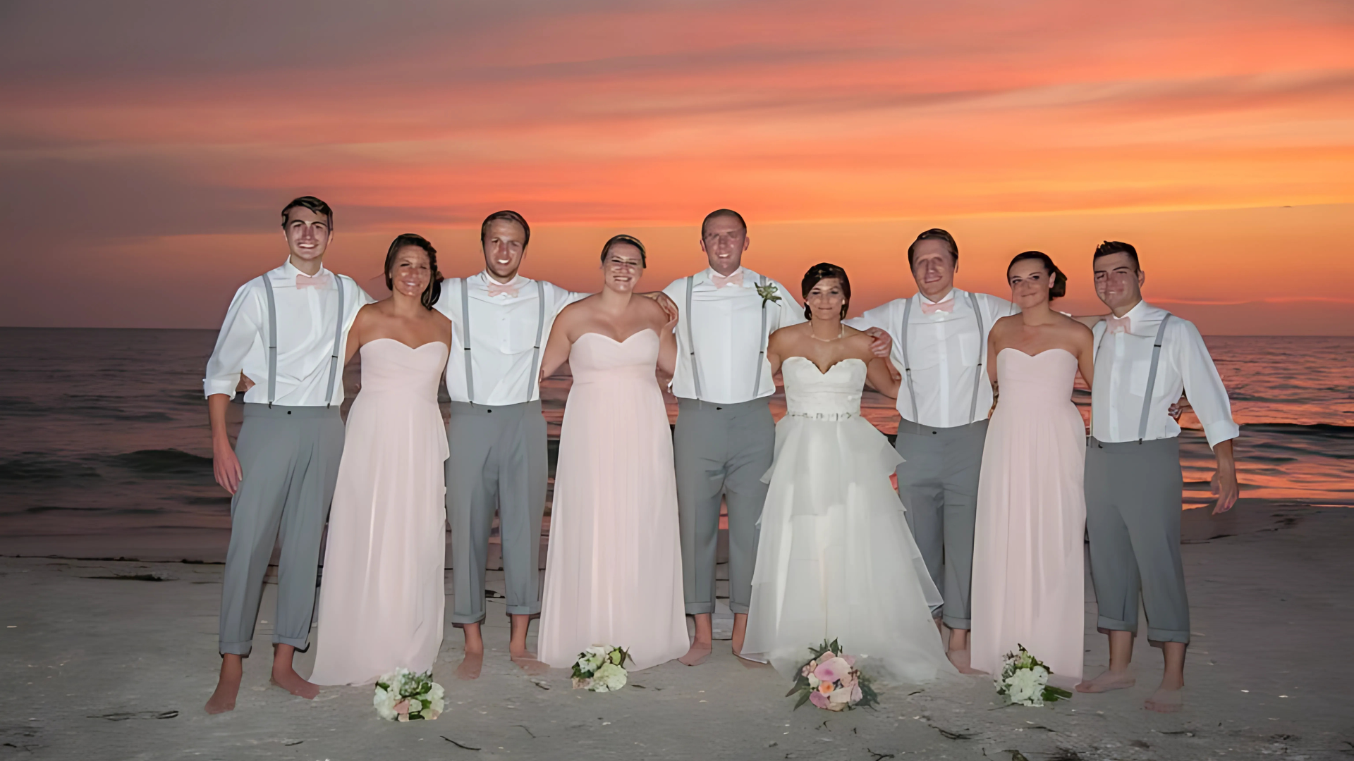Wedding party on beach at sunset. Bridesmaids in pink gowns, groomsmen in gray pants and suspenders.