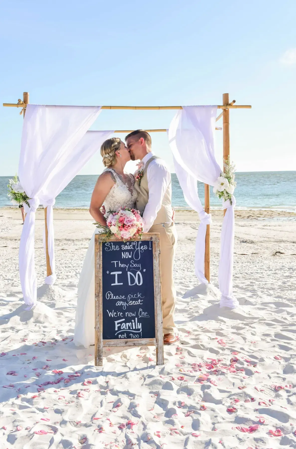 Couple kissing under wedding arch on beach; chalkboard sign says