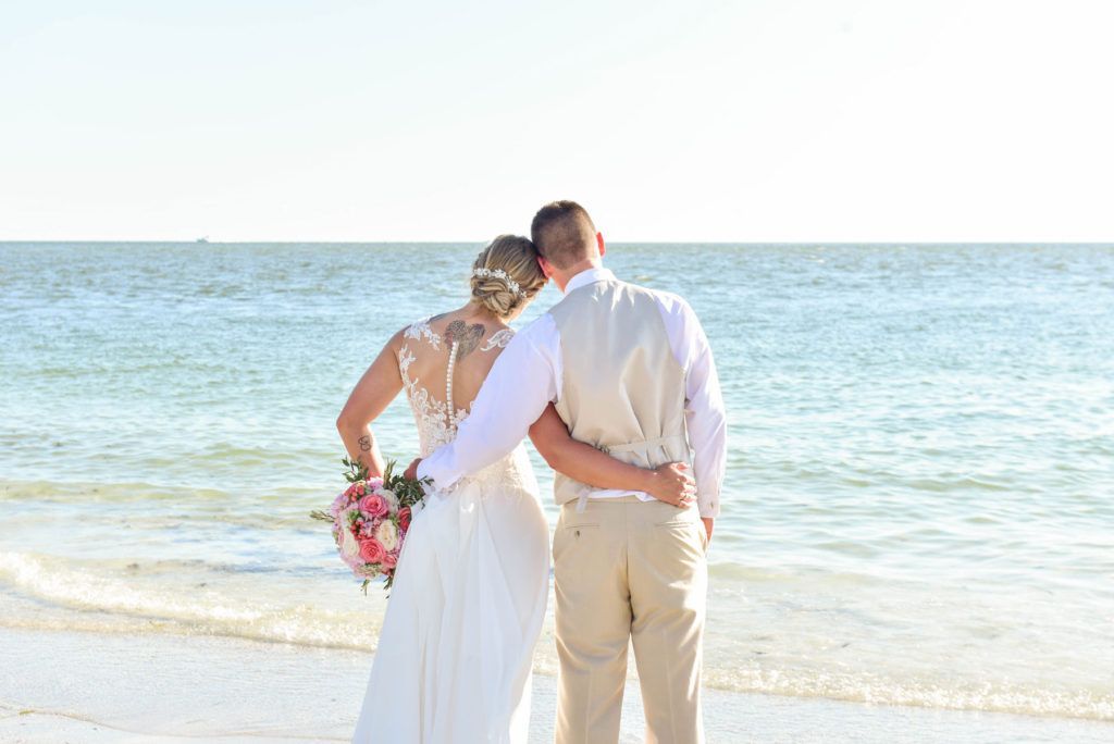 A bride and groom are standing on the beach looking at the ocean.