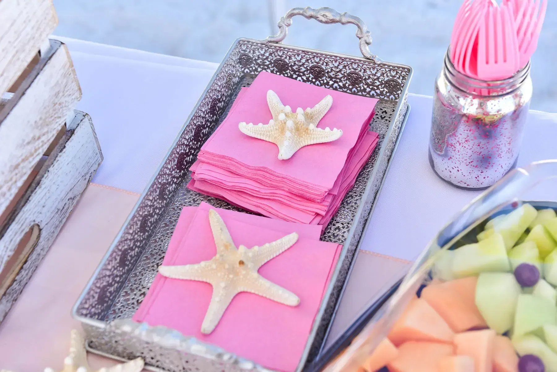 Tray with pink napkins and starfish, with pink forks in a glitter jar, and fruit.