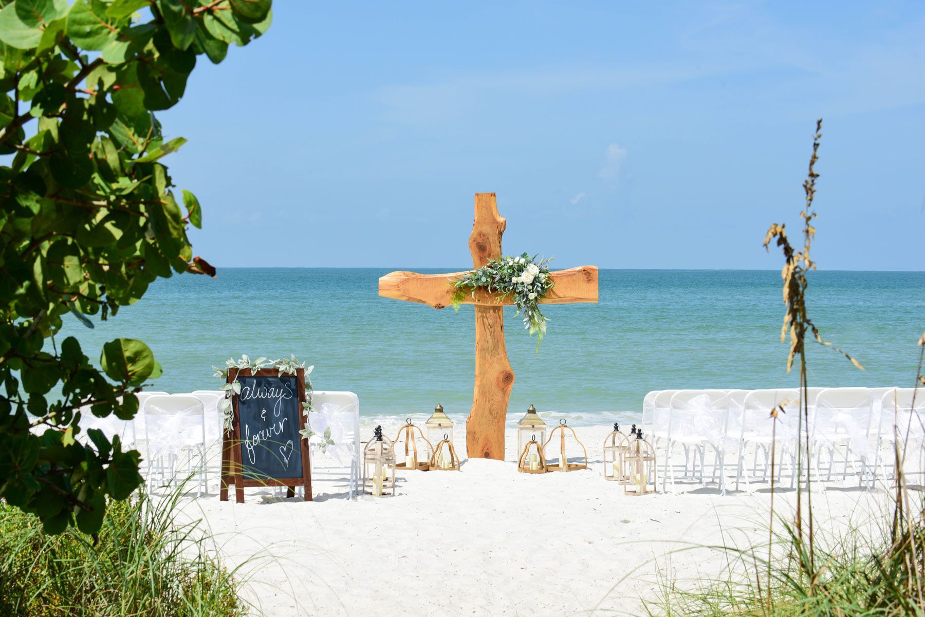 Beach wedding setup with a large wooden cross, chairs, and ocean backdrop.