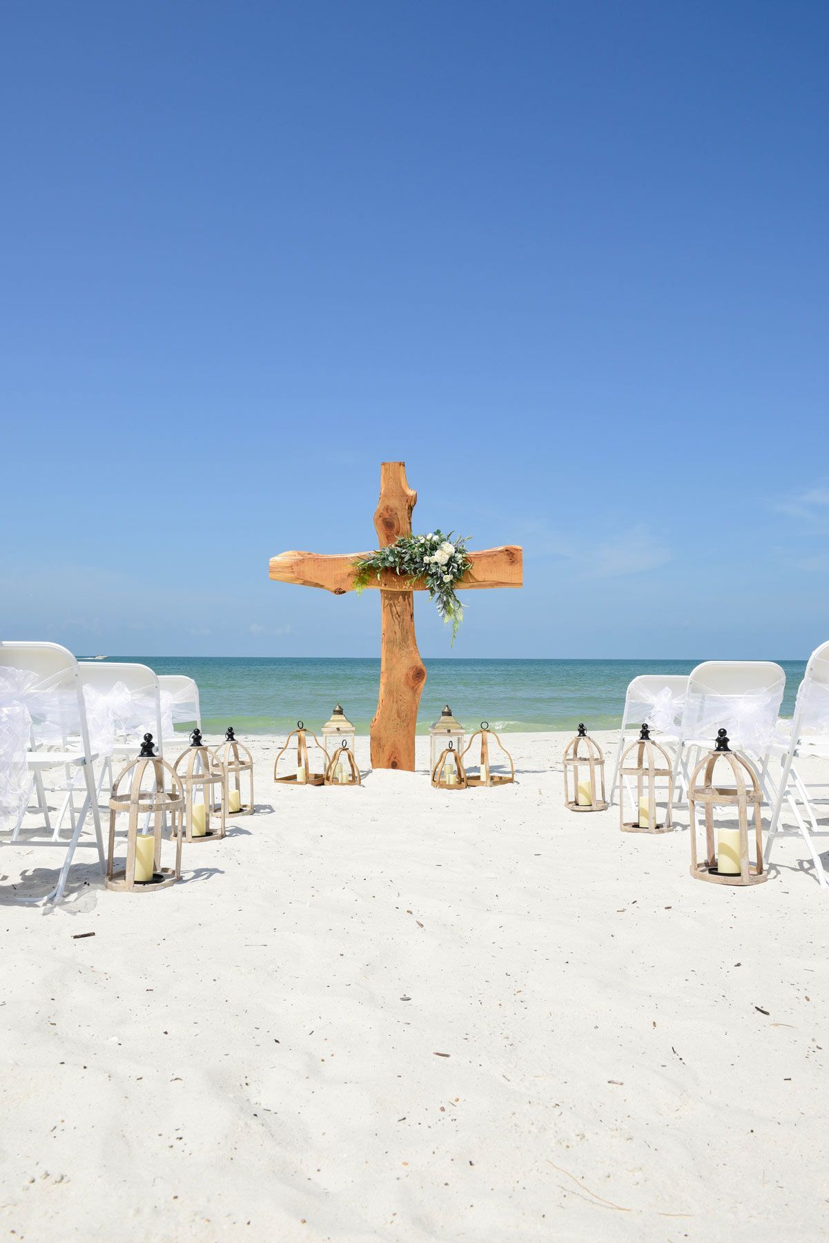 Beach wedding ceremony with wooden cross, lanterns, and chairs on white sand under a blue sky.