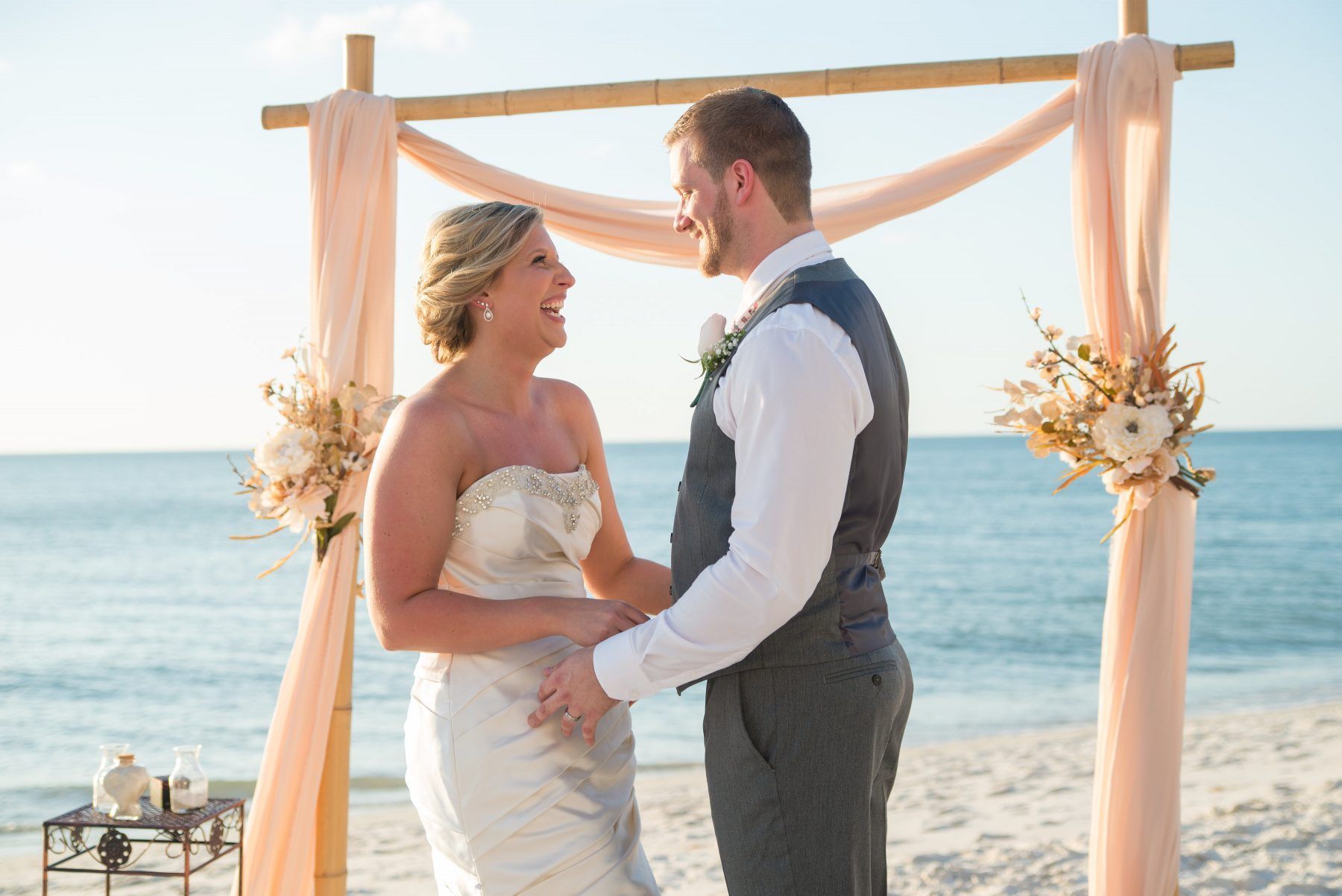 Bride and groom laugh, holding hands, on a beach during wedding ceremony. Bamboo arch with peach fabric and floral accents.