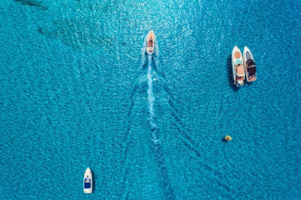 Boats on clear blue water. A motorboat creates a wake; others are docked.