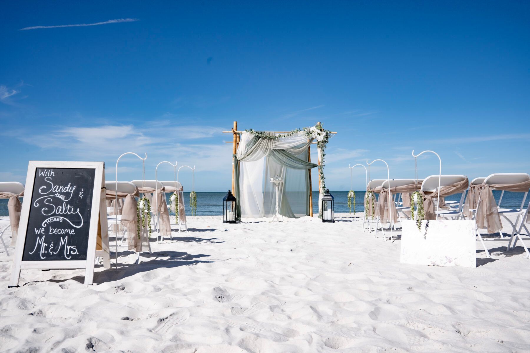 Beach wedding setup: white sand, blue sky, chairs, arch, chalkboard sign with text.