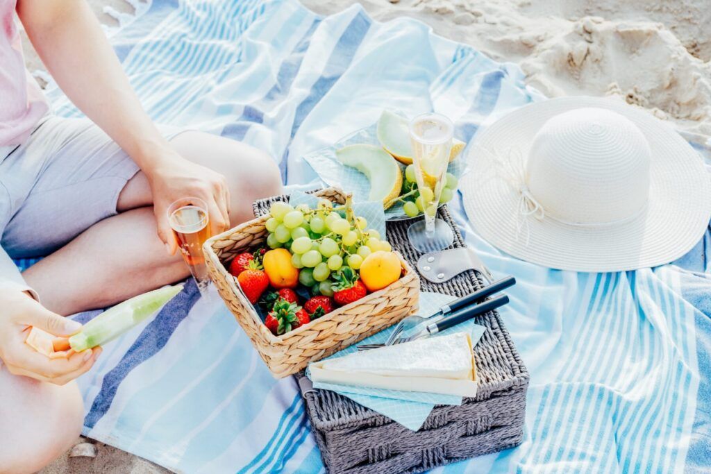 The cropped man sitting on the plaid with picnic food and drinks for two. Romantic date on the beach