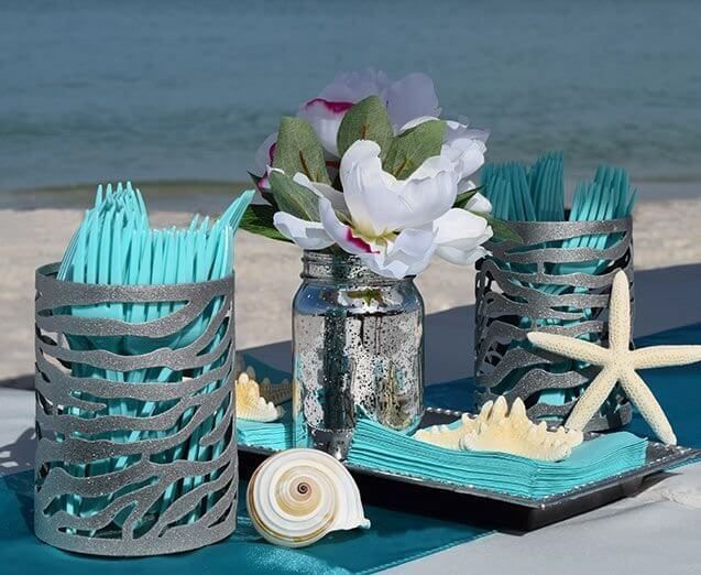 Beach-themed table setting with turquoise utensils, flowers, seashells, and a mirrored jar, against a blurred ocean backdrop.