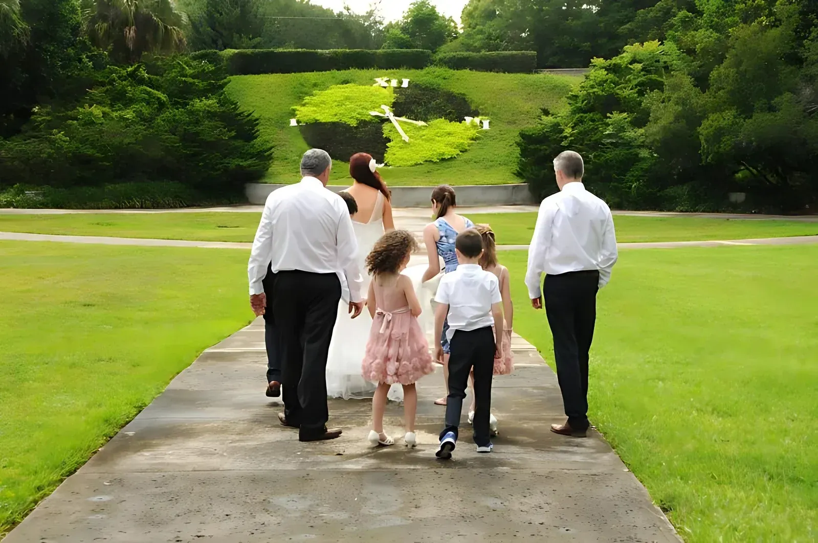 Wedding party walks on a pathway in a garden. Bride in white dress, children, and adults walk toward large floral clock.
