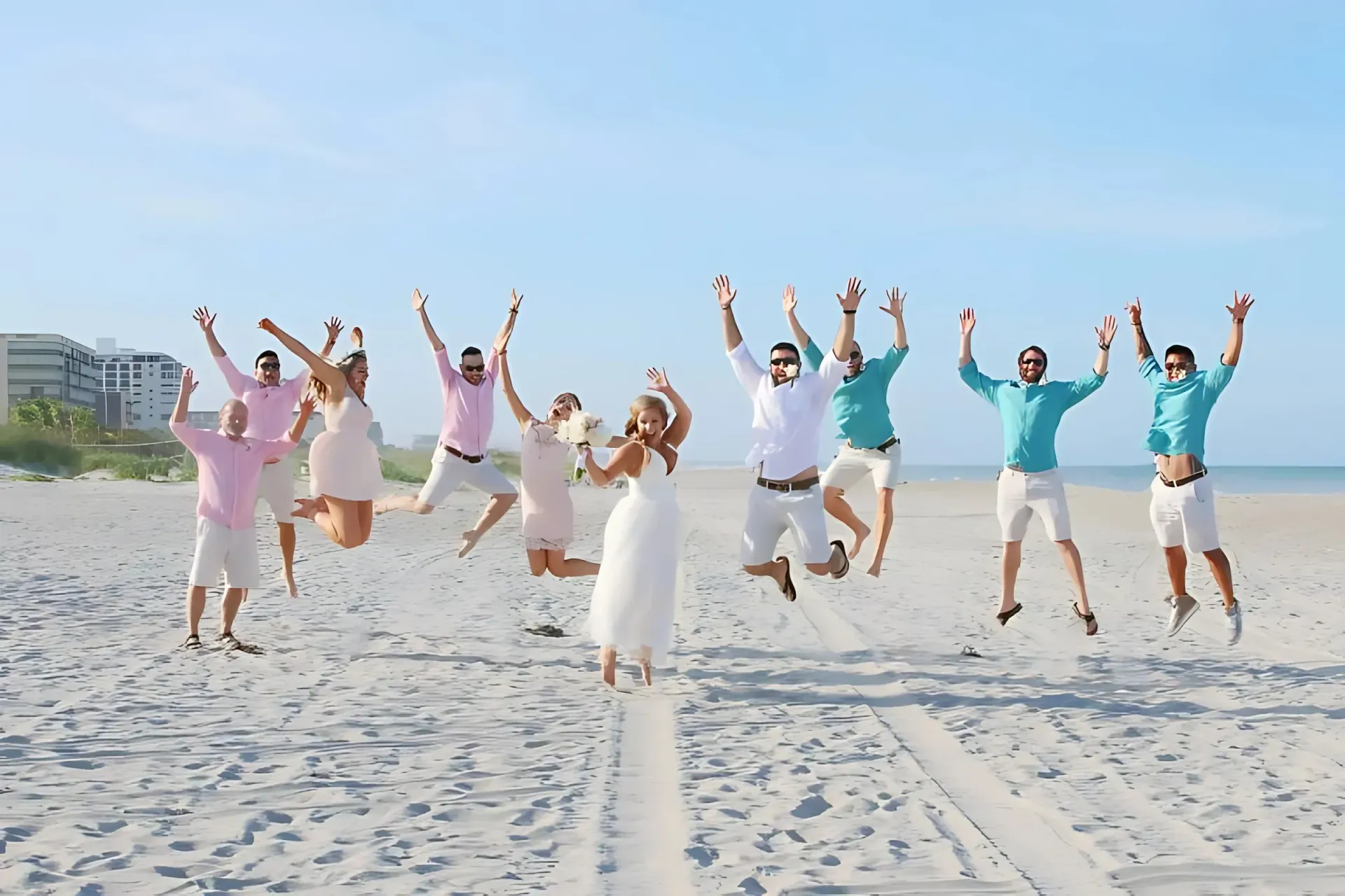Wedding party jumping on beach; men in white, pink, and teal shirts, bride in white dress.