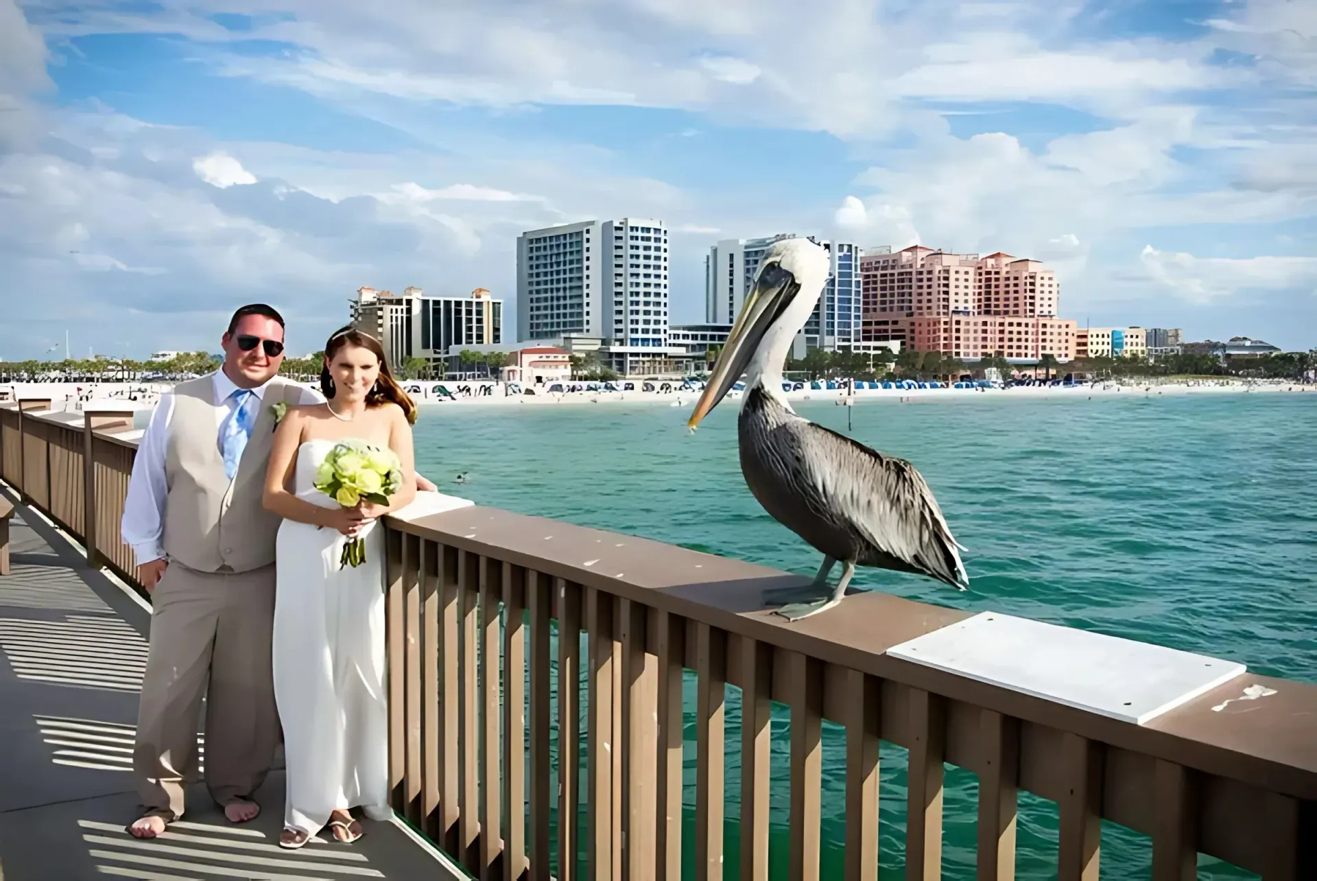 Newlyweds pose with a pelican on a pier overlooking a beach and city skyline.