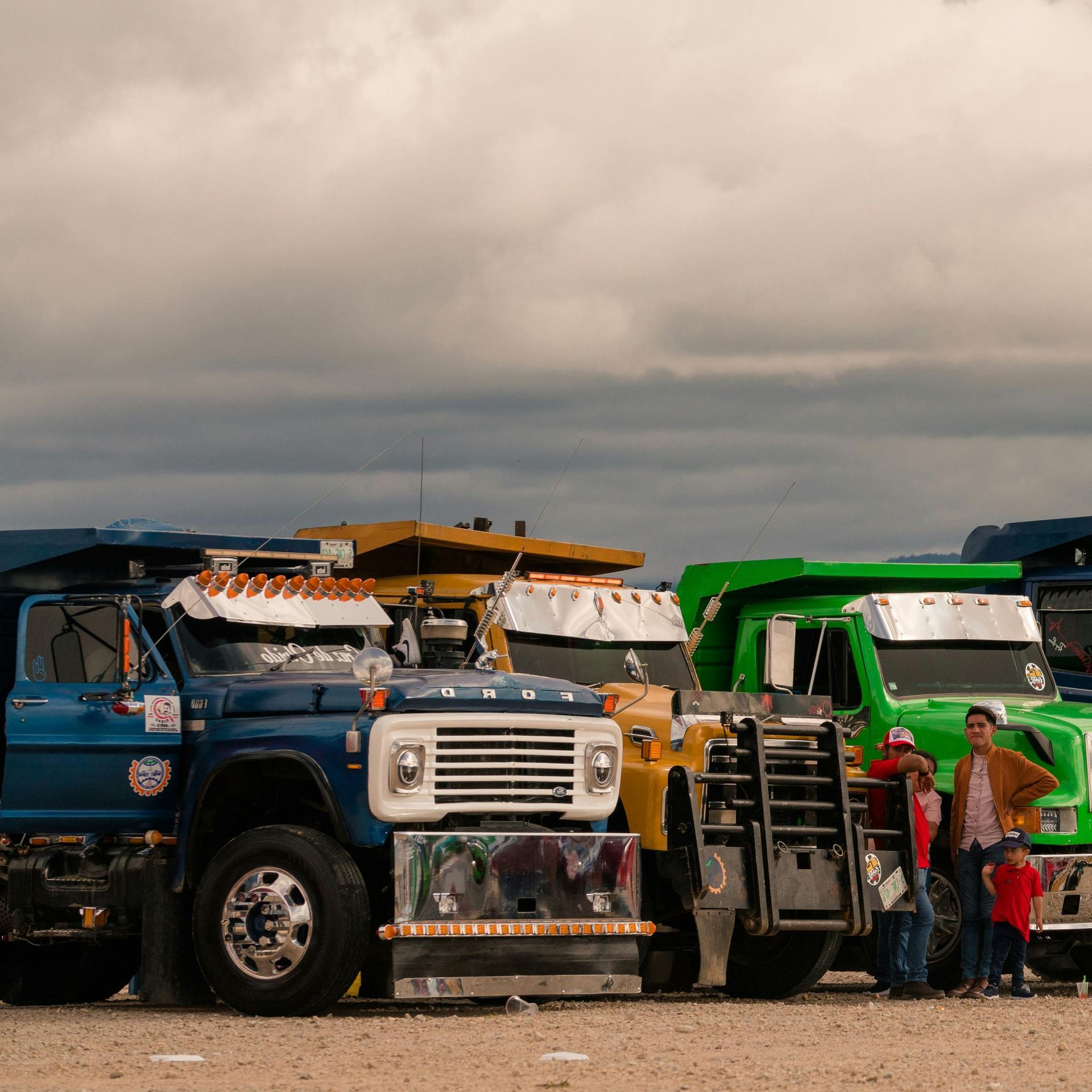 Colorful dump trucks parked on dirt under a cloudy sky, with people standing beside the green truck.