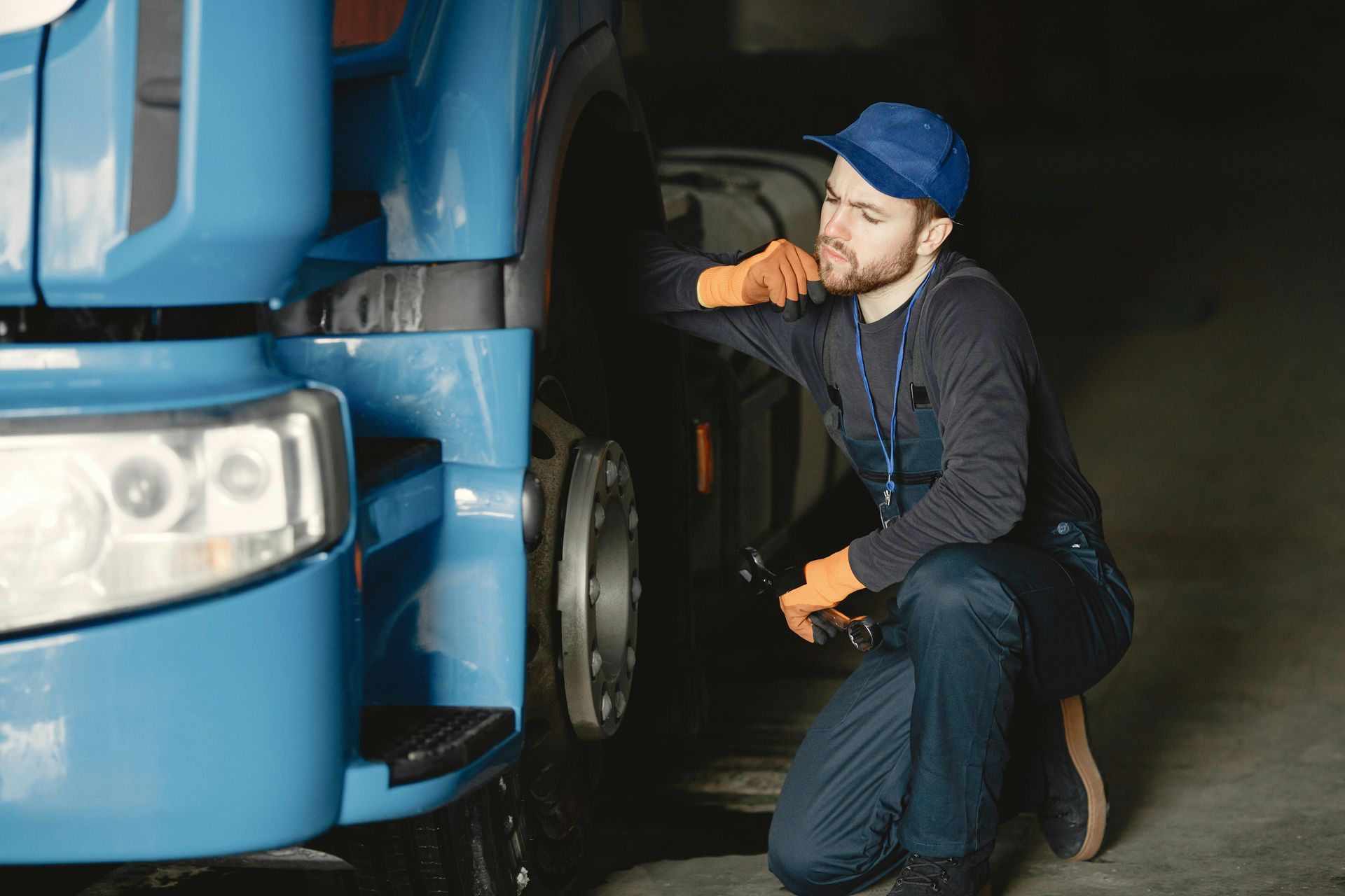 Mechanic in blue overalls inspecting a large blue truck's wheel. He wears a blue cap and orange gloves, looking thoughtful.