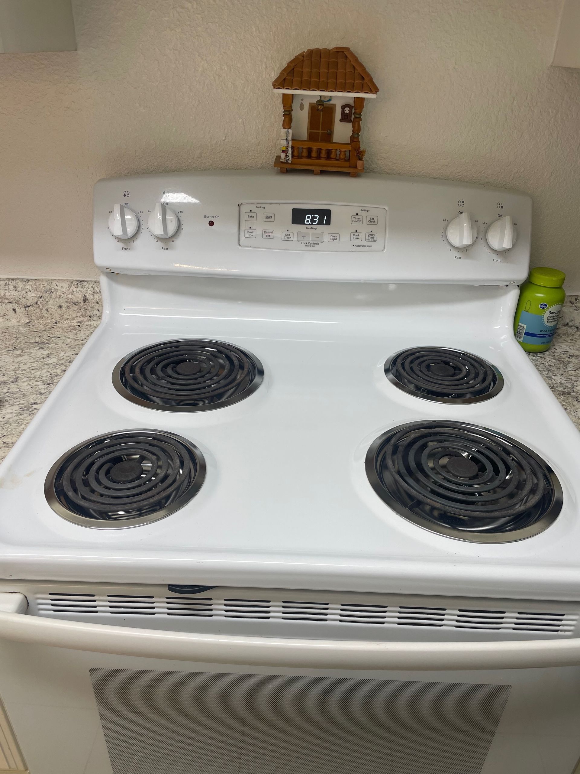 White electric stove with four black burners, a small decorative house on top, and a small container to the right.