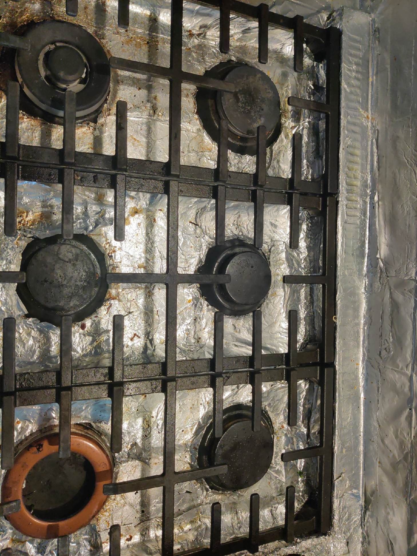 A top-down view of a dirty gas stovetop with five burners and black grates. It appears to be covered in aluminum foil.