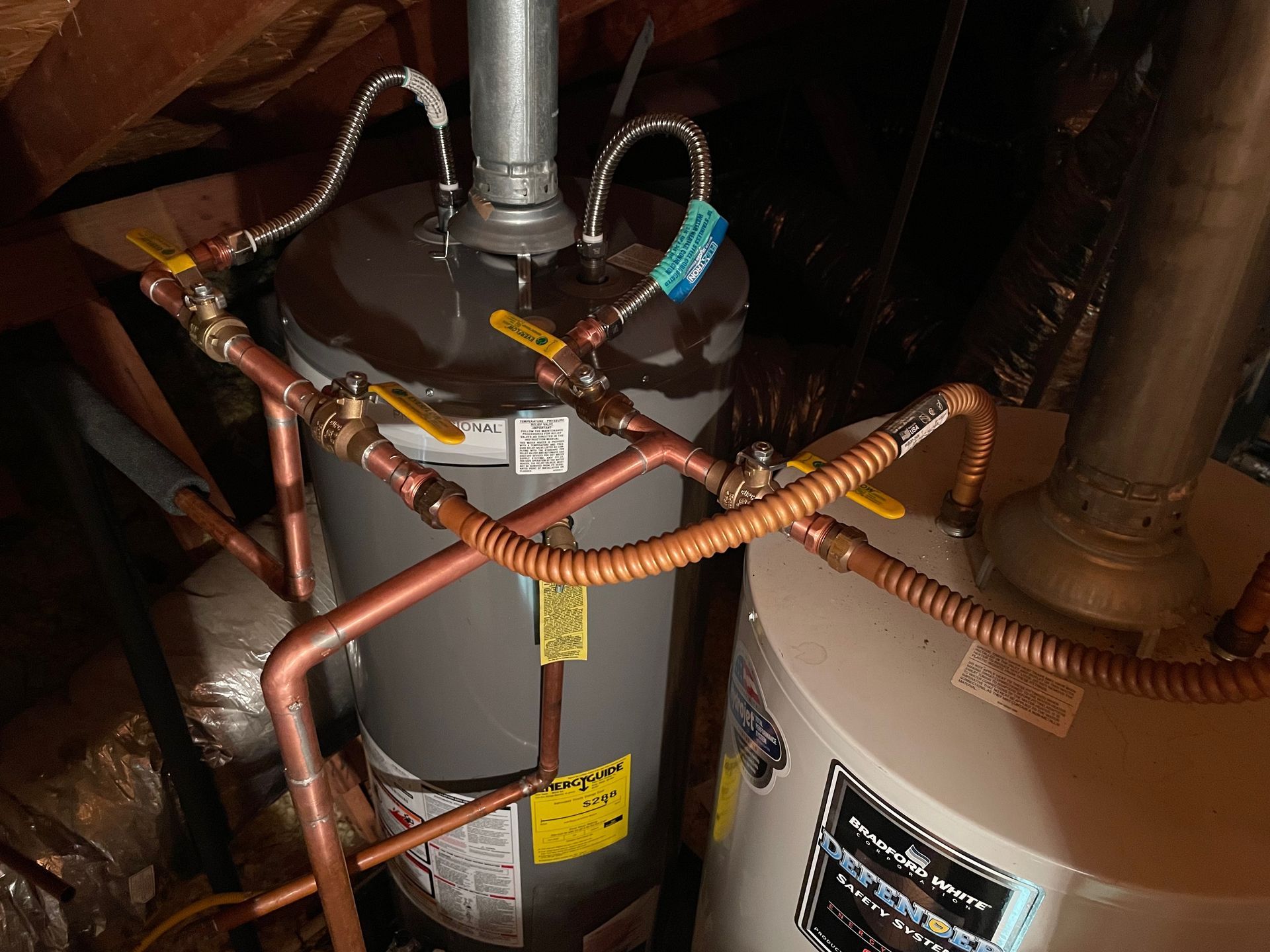 Two hot water heaters with copper pipes and yellow valve handles in a dark, attic-like space.