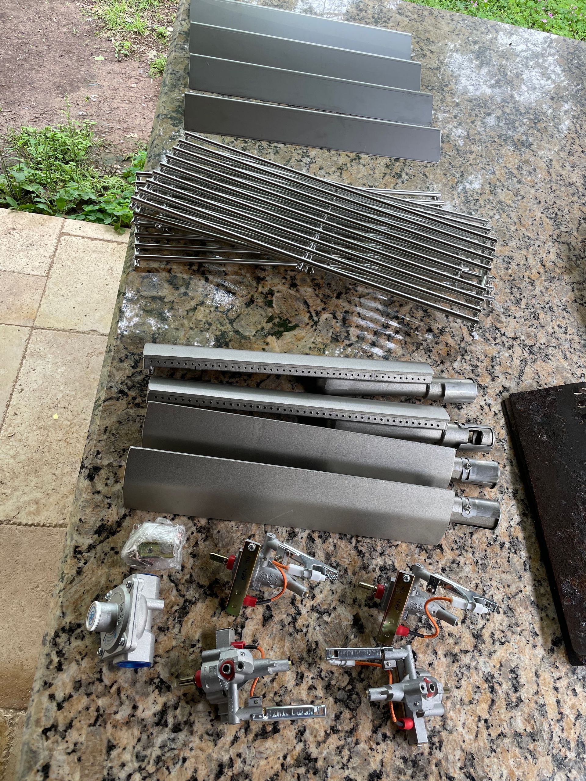Disassembled grill parts laid out on a granite surface: grill grates, burners, heat plates, and ignition components.