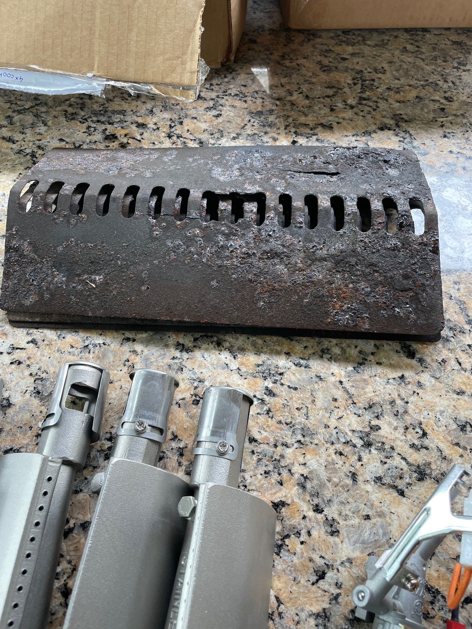 Close-up of a heavily corroded metal burner shield and several new gas grill burner tubes and control knobs.