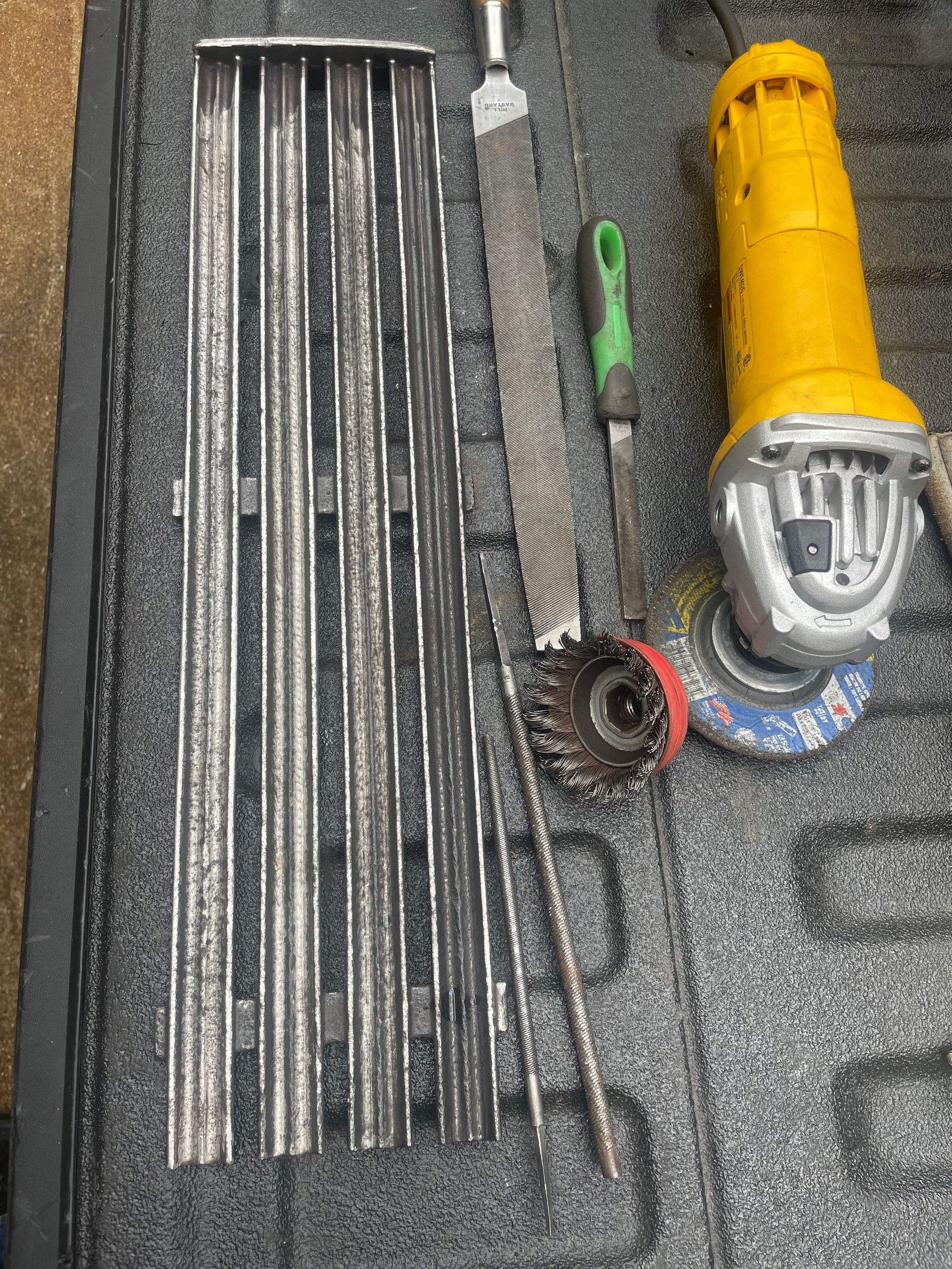 Grill grates and tools, including a wire brush, file, and angle grinder, on a textured surface, likely for cleaning or restoration.