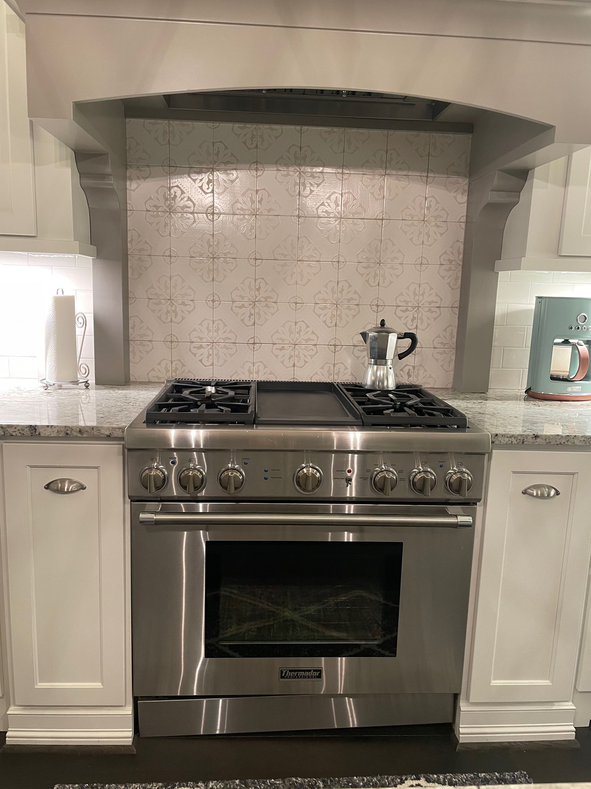 Stainless steel range in a kitchen with white cabinets and tile backsplash. A coffee pot sits on the stovetop.