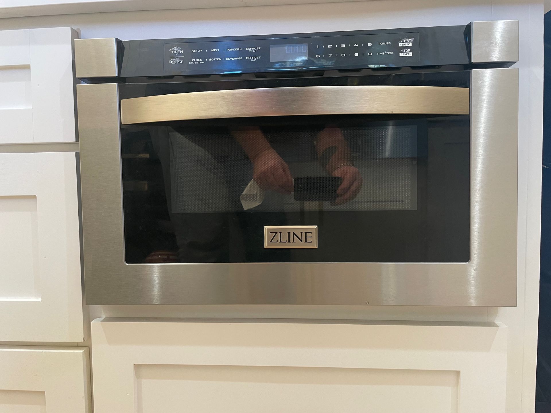 A stainless steel built-in microwave oven with black controls is installed in white kitchen cabinets. The microwave has a glass door and the brand name is ZLINE.