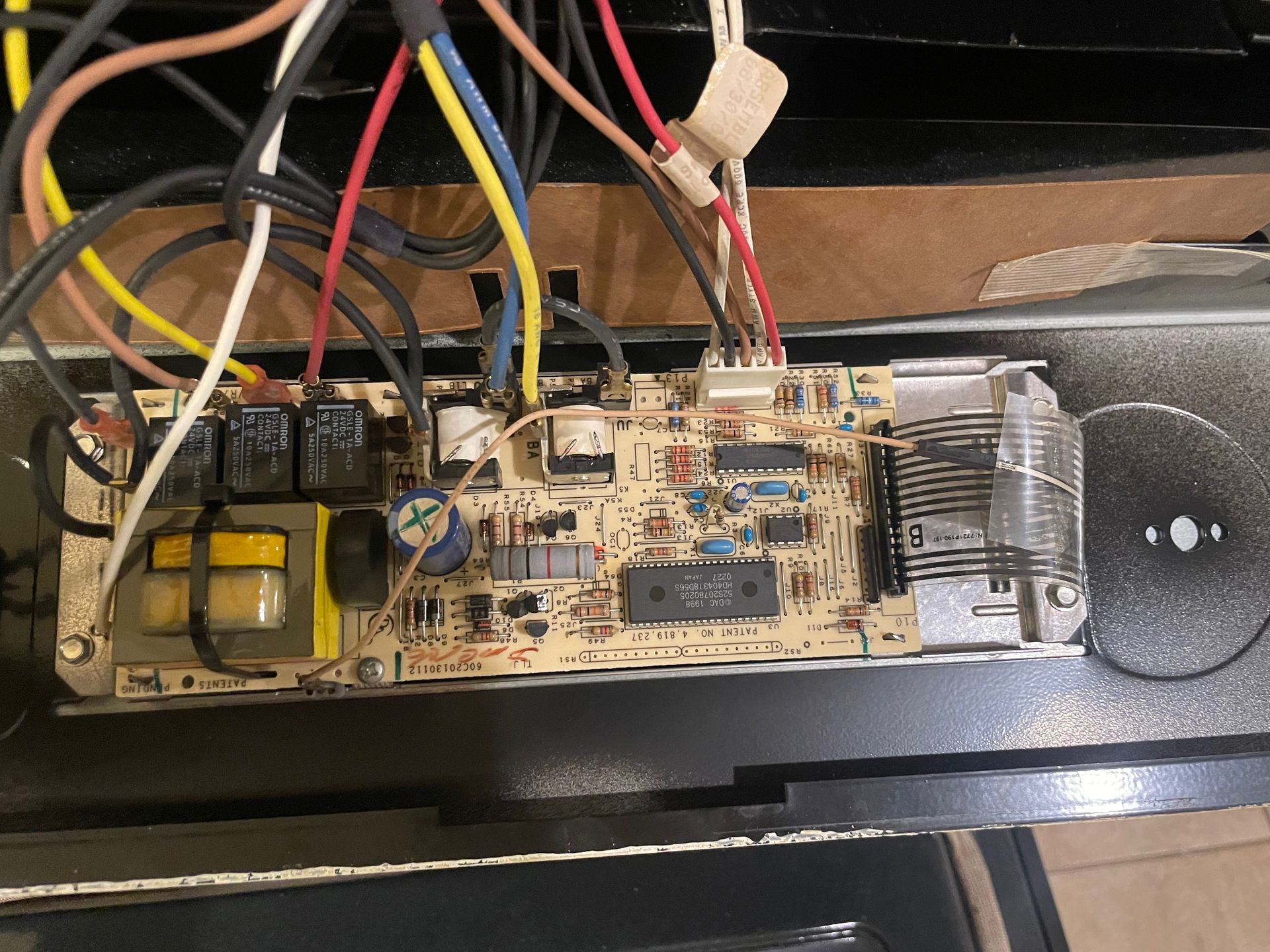 An oven control board with numerous wires of various colors attached to it, mounted inside a dark oven.