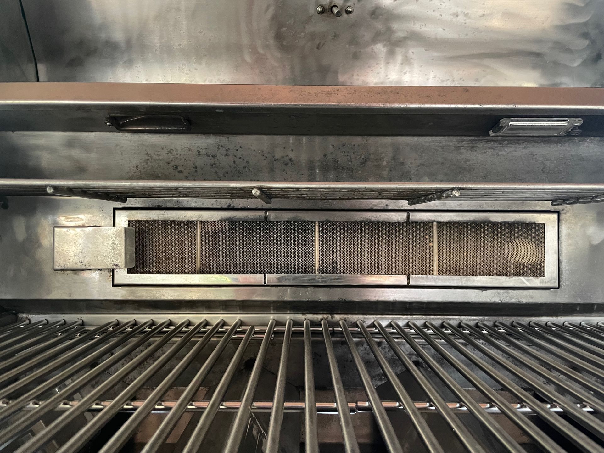 Inside of a grill, showing grates below and a burner above. The burner is a metal plate with small, rectangular openings.