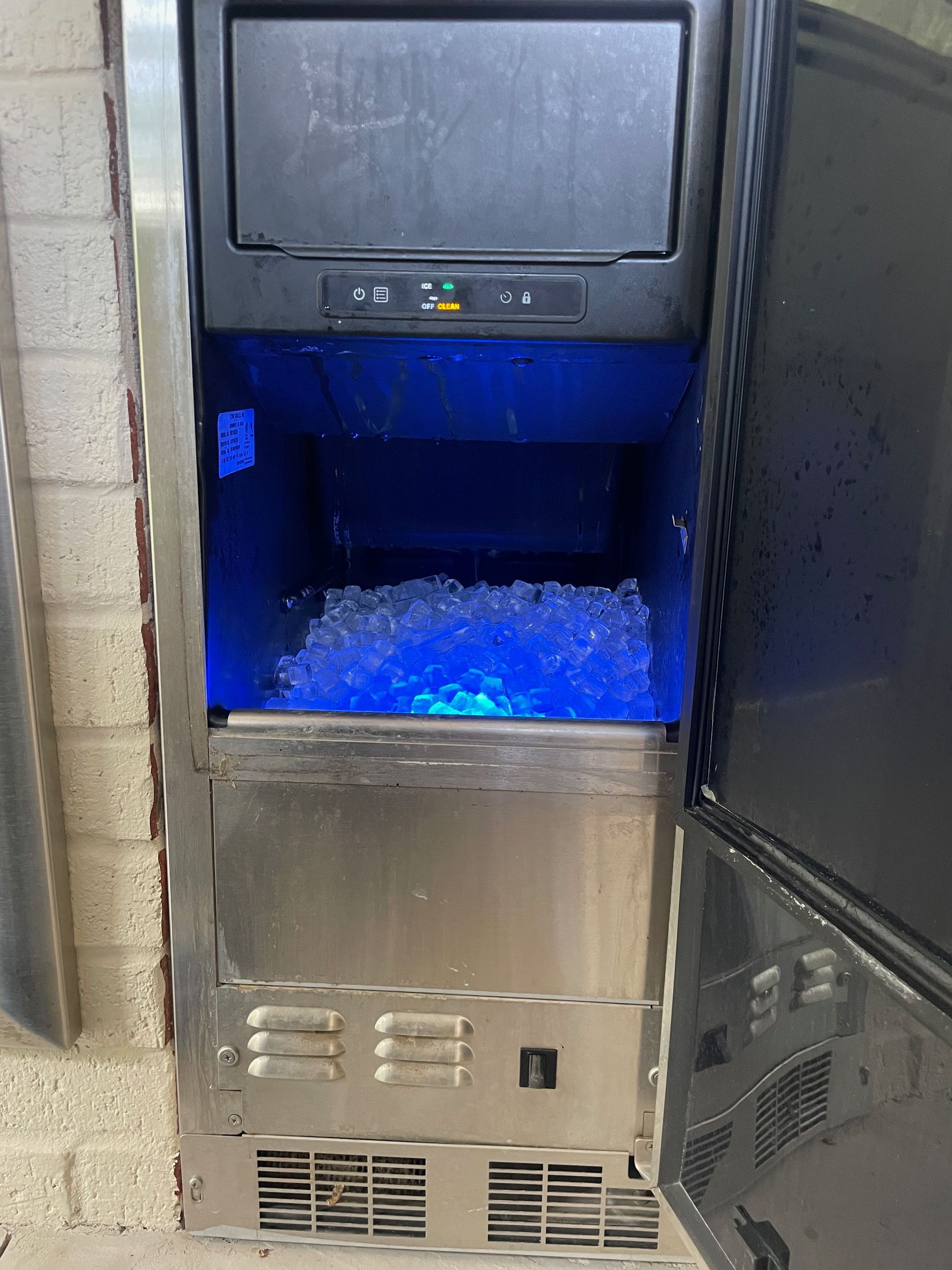 Ice machine with a door open revealing a bin of ice illuminated by blue light. Stainless steel exterior.