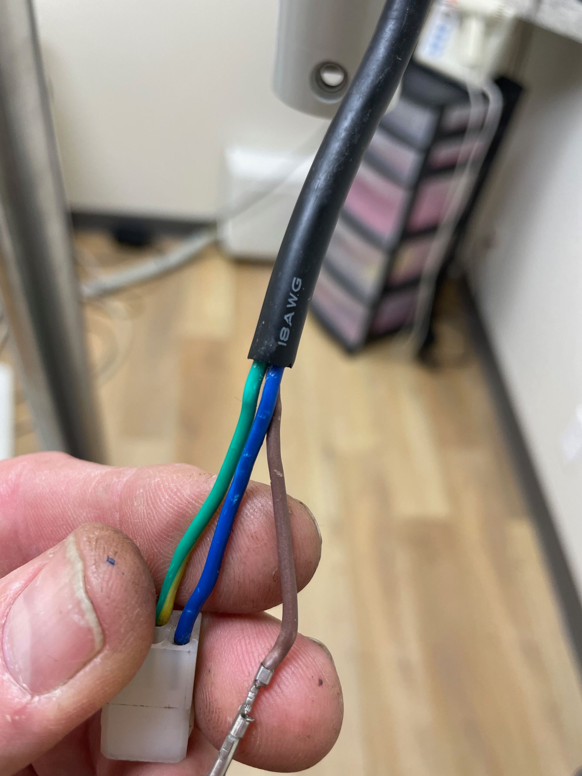 A hand holding a cut power cord with green, blue, and brown wires. The white connector is visible at the base of the wires.