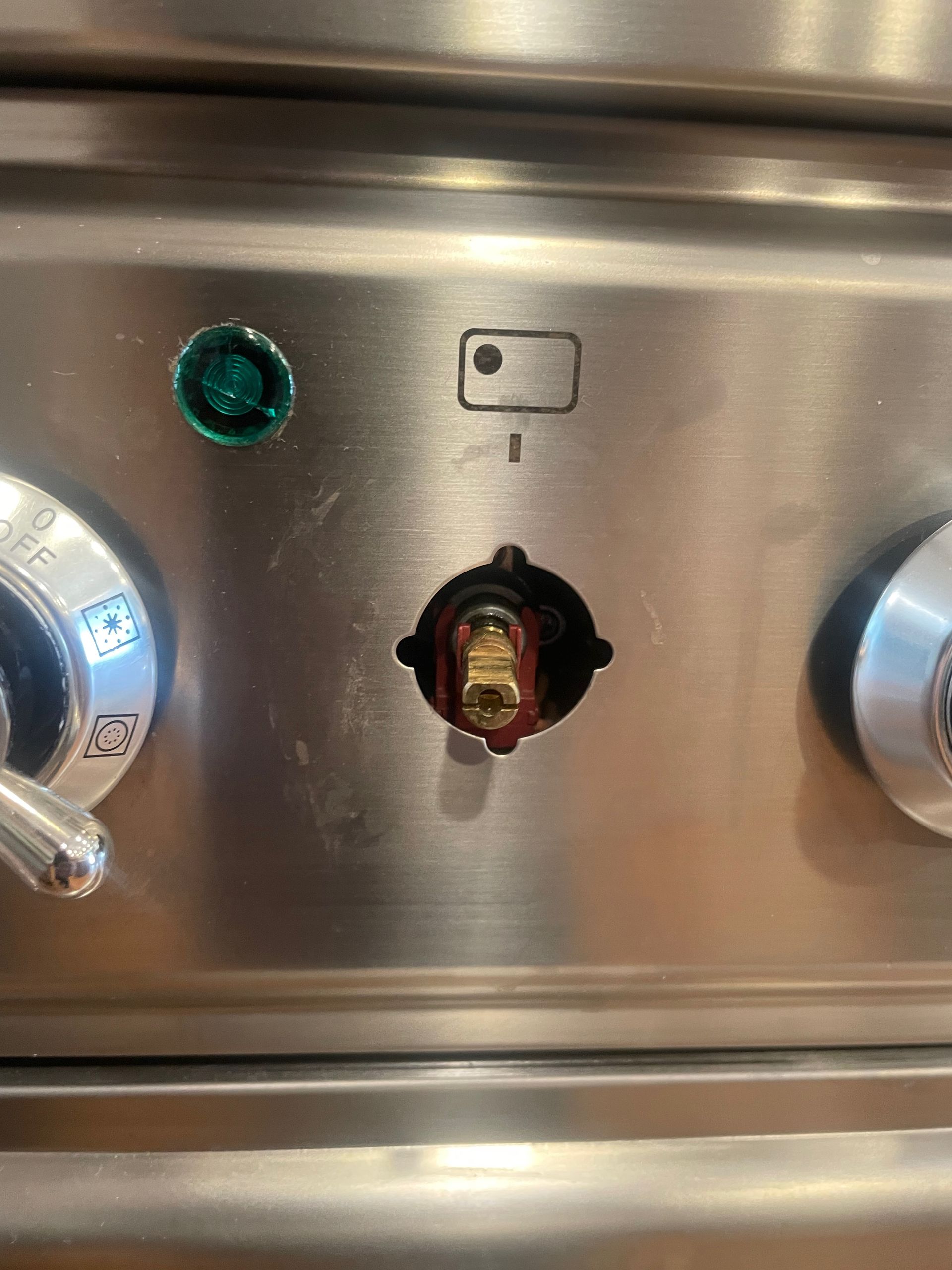 Close-up of a stainless steel oven control panel. A broken knob area is visible, with a green light and a small symbol above.