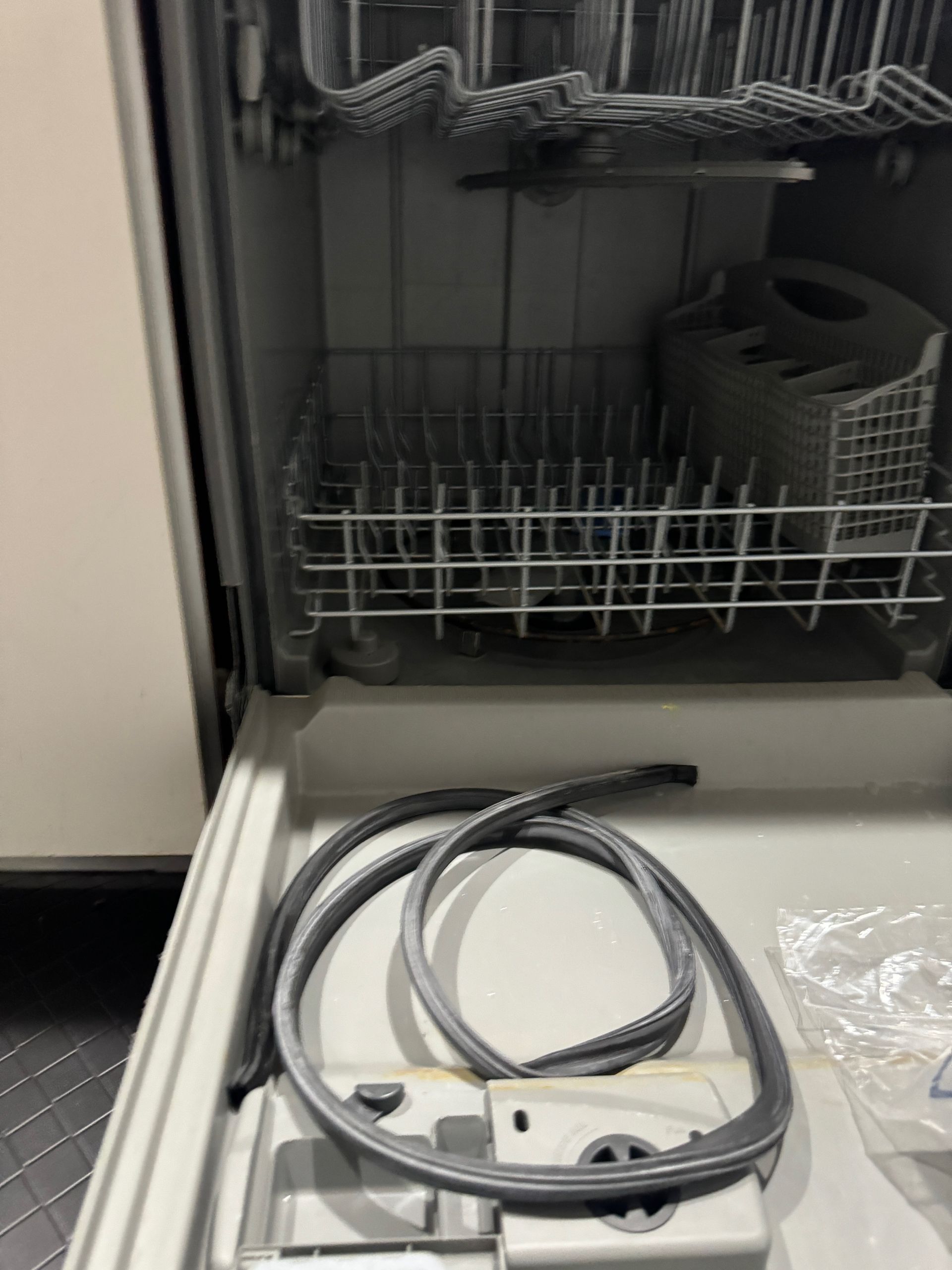 Open dishwasher with a grey, coiled rubber seal lying in the open door. Interior racks and utensil basket are visible.