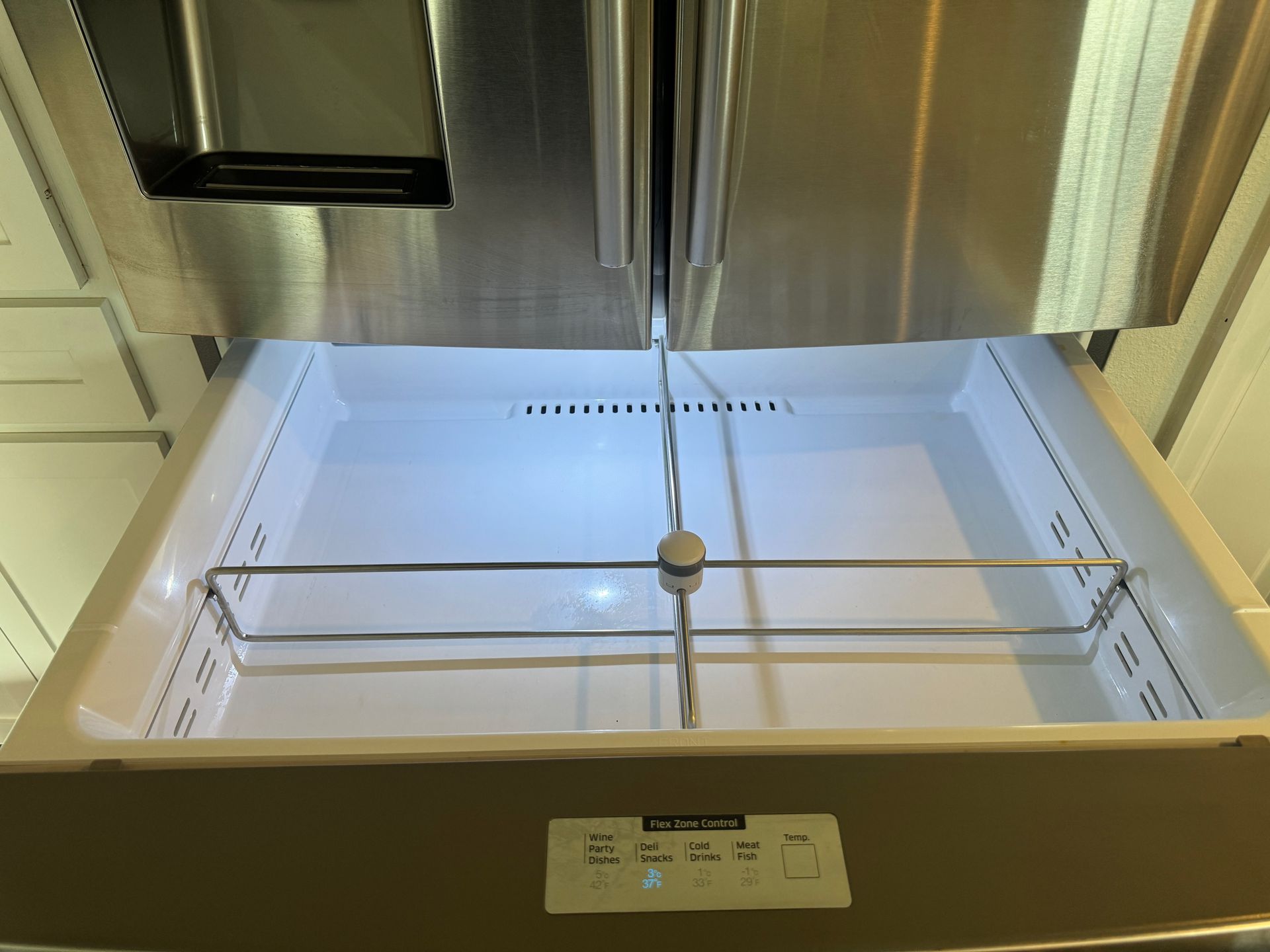An open refrigerator produce drawer with a wire rack, empty, and a control panel at the bottom. Stainless steel doors are above.
