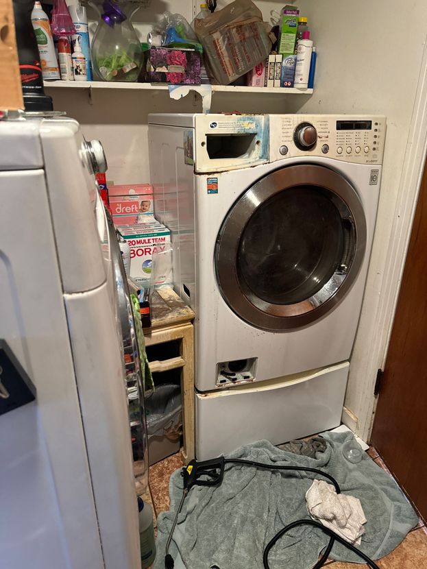 A white front-loading washing machine, possibly damaged, in a cluttered laundry area with cleaning supplies.