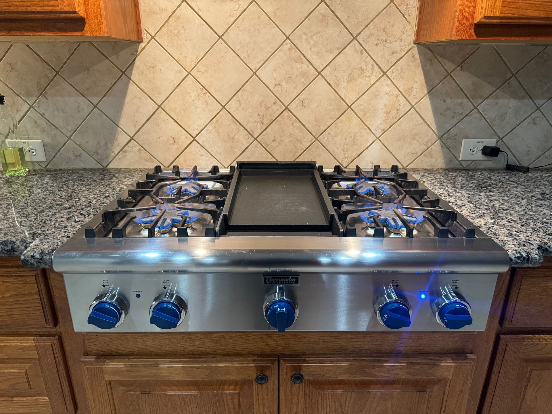 A stainless steel gas stove with blue flames and knobs is in a kitchen with granite countertops and a tiled backsplash.