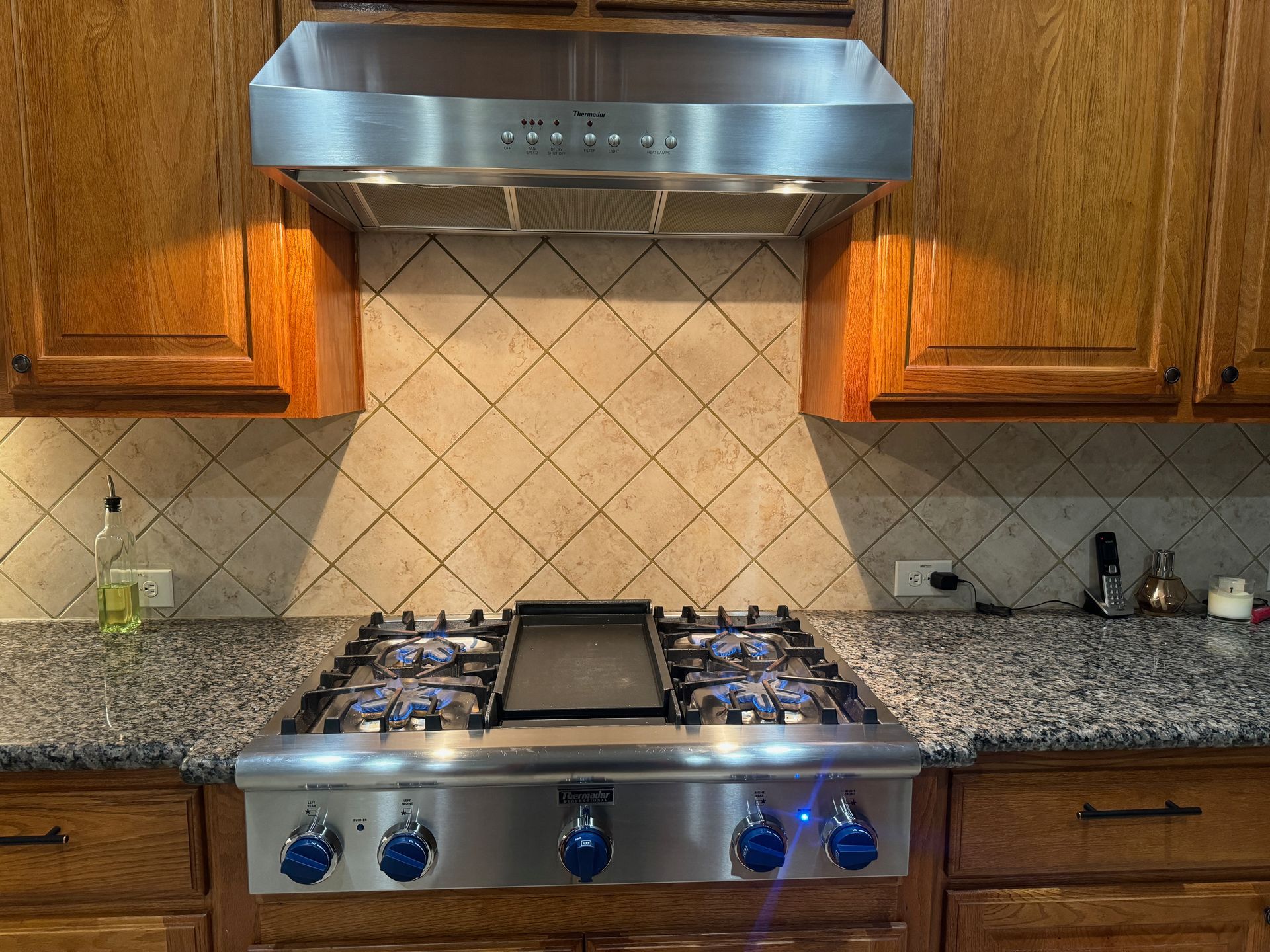 A stainless steel range with blue flames burning on the stove sits under a range hood in a kitchen with granite countertops and wood cabinets.